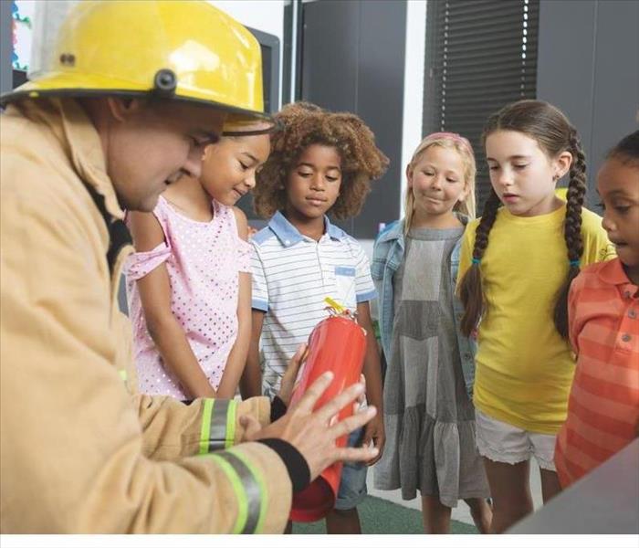 Rear view of Caucasian firefighter wearing fire helmet on his head and teaching about fire extinguishers to schoolchildren.